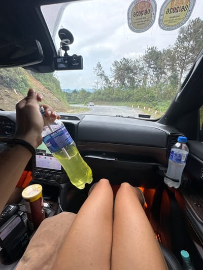 The image shows the interior of a car, possibly moving, in Quản Bạ Commune, Vietnam. The interior is cluttered with items including a half-empty bottle of yellow liquid, a bottle of water, a can of potato chips, and a smartphone. A hand is holding a bottle, seemingly with a straw inside. The car is on a road passing through a mountainous, forested area. Stickers are visible on the windshield. The legs of the person taking the photo are also visible in the frame. A dashboard camera is mounted on the windshield.