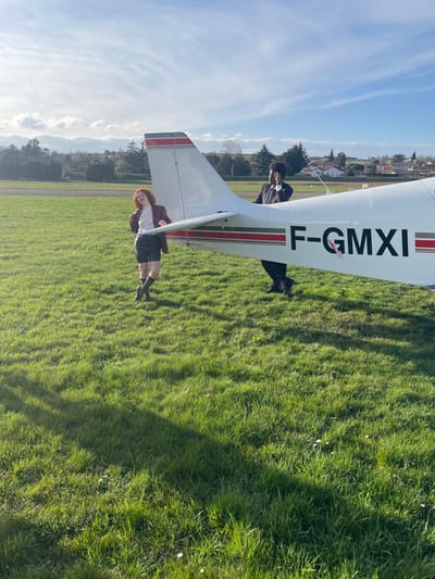 The scene depicts an outdoor setting on a grassy airfield, identified as Aérodrome de Vienne - Reventin (LFHV) in Reventin-Vaugris, France, under a partially cloudy blue sky. A white single-engine light aircraft, bearing the registration F-GMXI with horizontal red and black stripes, is centrally positioned on the grass.
Two individuals are present near the aircraft. To the left of the aircraft's tail, a person with red, curly hair is standing, clad in a white shirt, dark leather shorts, a dark red blazer, dark brown knee-high socks, and dark brown ankle boots. This individual's right arm is slightly raised, and the left hand is near the hip, with their gaze directed towards the camera.
Partially obscured behind the right side of the aircraft's fuselage, a second person with dark, textured hair is visible. This individual is dressed in a dark suit, white shirt, and dark tie. Their right hand is raised near the ear, and their gaze is directed away from the camera, towards the background.
The immediate environment consists of vibrant green grass with scattered white wildflowers. In the mid-ground, a paved strip (likely a runway or taxiway) is visible. Further in the background, residential buildings with red-tiled roofs and numerous trees, both deciduous and evergreen, are present under a slightly hazy horizon. Long shadows are cast across the grass from the left, indicating a low sun angle.