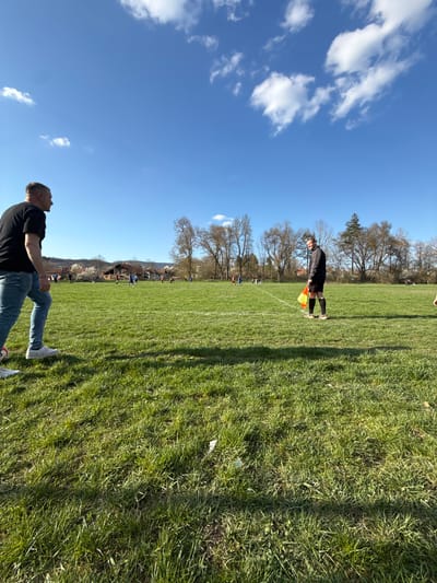 A sunny outdoor scene captures a soccer match in Dushevo, Bulgaria. The blue sky is dotted with scattered clouds. Two men are visible on the sidelines: one is standing with a flag in hand, clad in a black uniform, while the other is seated, wearing a black shirt and blue jeans. The soccer field is well-maintained with green grass and white lines. In the background, trees and houses are visible, suggesting a rural or suburban environment. The players are visible on the field.