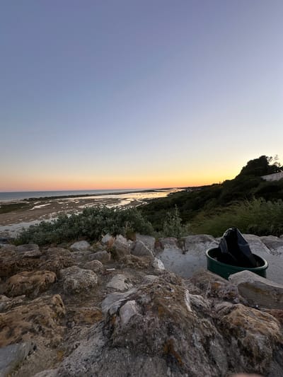 The image shows a landscape at the mouth of the Guadiana River in Vila Real de Santo António, Portugal, during dusk or dawn. The sky transitions from blue to orange and yellow near the horizon. The river, partially exposed with sandbanks, is visible in the middle ground. In the foreground, a rough stone wall is topped with vegetation and a green trash can containing a black bag.