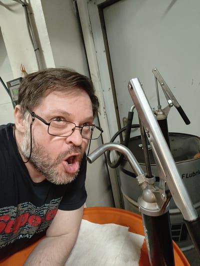 The image is a selfie taken by a Caucasian man with short brown hair and a beard. He is wearing glasses and a black t-shirt with red lettering. His mouth is open and he is looking at the camera with a surprised expression. He is positioned in front of a large metal barrel and a grease gun. The background appears to be an industrial or workshop setting, with pipes and electrical conduits visible on the wall. The image does not contain any information about the specific location within the city of Ягуново, Russia.