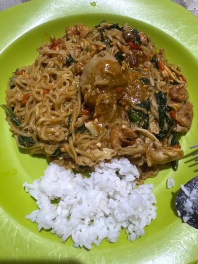 An overhead shot shows a bright green plate of food. In the upper portion of the plate, a generous serving of instant noodles is mixed with pieces of chicken, leafy greens, and what appears to be chili. The noodles are coated in a thick, savory sauce. To the left, a mound of white rice sits in the lower half of the plate. A fork is visible on the right side of the plate, partially obscured by a dark-colored spoon with some food residue. The food is presented on a patterned tablecloth. This image was captured in Kalijaten, Indonesia.