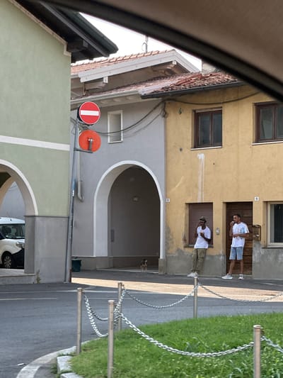 Two individuals are captured in a medium shot, standing on a street outside a building. The individual on the left, dressed in a white t-shirt and blue shorts, is holding a white object to their mouth. The person on the right wears a white t-shirt and khaki pants, also with a white object near their mouth. They appear to be in an urban or semi-urban setting, possibly in Rudiano, Italy, indicated by the architectural style of the buildings and the presence of chain barriers alongside a grassy area and a paved road. The time of day appears to be daytime, with bright sunlight casting shadows. One building features visible house numbers "15" and "13". Red and orange circular signs are mounted on a pole. In the background, parts of other buildings and a white vehicle are visible. The scene suggests a casual moment, with the individuals engaged in an activity that appears to involve eating or drinking.