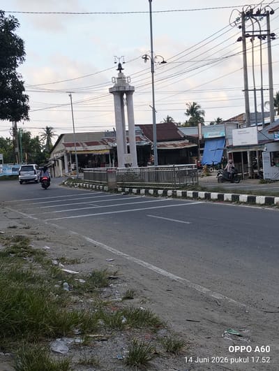 The image depicts a street scene in Ulee Pulo, Indonesia, with a prominent white monument in the mid-ground, topped with scales of justice. The monument stands at an intersection with a paved road. To the left of the monument, a silver SUV is driving away from the camera, and a person on a motorcycle is also moving away from the viewer. Shops with corrugated tin roofs line the right side of the road, with various signs visible, including "MYVIDIA GROSIR" and "RAHFA CELL." A blue tarp is draped over a structure behind one of the shops. A motorbike with a rider is parked in front of one of the shops. The sky is overcast. The foreground shows a grassy verge littered with trash, including plastic bottles and wrappers. Power lines and utility poles are visible throughout the scene, indicating an urban or semi-urban environment. The bottom right of the image contains a timestamp: "OPPO A60 14 Juni 2026 pukul 17.52".