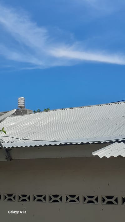 This is a photograph taken in Bireuenmeunasahreuleut, Indonesia, featuring a partially visible building with a corrugated metal roof. A water tank sits atop a concrete structure on the left side of the roof. The sky is blue with wispy white clouds. The bottom of the image shows a section of the building's exterior wall with decorative ventilation blocks. The camera's timestamp "Galaxy A13" is visible in the lower left corner.