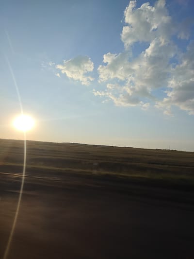 The image captures a vast, rural landscape under a clear blue sky with scattered white clouds. The sun, a bright orb, is positioned in the upper left quadrant, casting a strong lens flare and illuminating the scene. The foreground is dominated by a dark, blurred area, likely indicating movement from a vehicle. This is followed by a wide expanse of dry, golden-brown vegetation that stretches to the horizon. The horizon is relatively flat, with a few sparse, dark vertical structures, possibly utility poles or distant trees, visible against the sky. The overall impression is of an open, agricultural or steppe environment, likely in the vicinity of Жаңаауыл, Kazakhstan, during daylight hours.