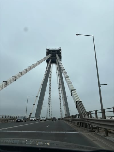 The image captures a forward-facing view from inside a moving vehicle on the Agigea Bridge over the Danube-Black Sea Canal in Agigea, Romania, under an overcast sky. The prominent central feature is a large, grey concrete pylon forming an 'A' shape, topped by a rectangular steel observation or maintenance platform. White cable stays, bundled and secured with black bands, extend diagonally from the pylon to the bridge deck. The bridge deck, featuring two lanes of traffic, stretches into the distance, with approximately ten passenger vehicles, including cars and one white van, visible traveling away from the camera. A metal guardrail runs along the right side of the bridge deck, with multiple grey streetlights spaced along the bridge. A tall streetlight stands on the right foreground. Reflections from the vehicle's dashboard are visible at the bottom of the frame. Small dark specks are discernible against the grey sky, and a circular dark smudge appears in the upper left quadrant of the image. No individuals are distinctly visible.
