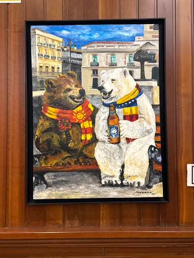 This is a painting of two anthropomorphic bears sitting on a park bench in front of buildings that appear to be in a European city, possibly Spain. The bear on the left is brown and wearing a scarf with a sun emblem. The bear on the right is white and holding a bottle of Polar brand beer. Both bears are smiling. The background shows buildings with balconies and windows, and a lamppost. The painting is framed in black and hangs on a wood-paneled wall. The signature on the painting reads "Morans '20". The location is not specified within the image itself.