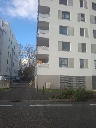 The image depicts an urban scene in Saint-Martin-d'Hères, France, on a clear day. The foreground shows a dark asphalt street with a single white horizontal line painted on its surface. A concrete curb separates the street from a paved sidewalk. To the right, a multi-story, light grey residential building occupies the majority of the frame, featuring multiple balconies with light-colored railings on its left facade and numerous windows, some equipped with closed or partially open grey shutters. The building's ground floor has a darker grey base with a metal grate and a square panel embedded into the wall. Three dark bollards are positioned on the sidewalk adjacent to this building. A patch of green vegetation and a larger bush are situated between the street and the building's base. To the left, a second multi-story, light grey building is partially visible, showing multiple window arrays. Between the two buildings, a paved area contains several parked vehicles, including white vans and dark cars. Bare deciduous trees are visible beyond the paved area. The sky above is clear blue with scattered white cumulus clouds. No individuals are visible.