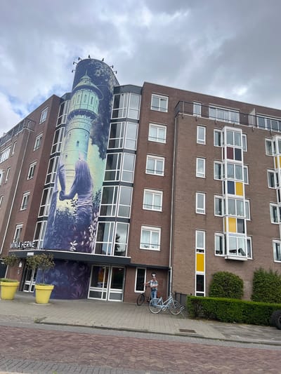 This media file captures a view of a multi-story brick building with a large mural on its side. The mural depicts a water tower surrounded by trees and a figure, possibly a woman, interacting with what appears to be a small animal. The upper sections of the building feature numerous windows, some with colored panes in yellow, blue, and dark red, creating a mosaic effect. On the ground level, near the entrance, a man is standing beside two light blue bicycles. The scene appears to be set on a cloudy day, with the sky taking up a significant portion of the frame. In front of the building, there is a paved sidewalk and a street made of brick pavers. Two large yellow planters with small trees are placed near the entrance. Visible text on the building includes "WINIA HERNE" printed horizontally. The location is identified as Leeuwarden, Netherlands.