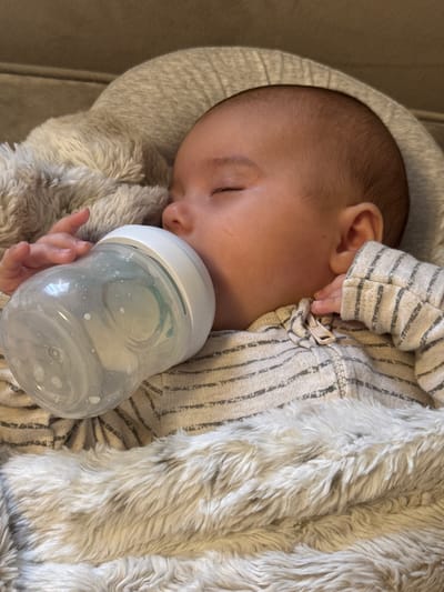 The image shows a close-up, slightly angled shot of a baby sleeping and drinking from a clear baby bottle. The baby is lying down, wrapped in a soft, fuzzy, light-colored blanket and a striped onesie. The baby's eyes are closed, and their hand is holding the bottle. The background appears to be a neutral-colored surface, possibly a couch or bed. The lighting is soft and natural. There is no visible indication of the city of Oldham, United Kingdom, in this image.