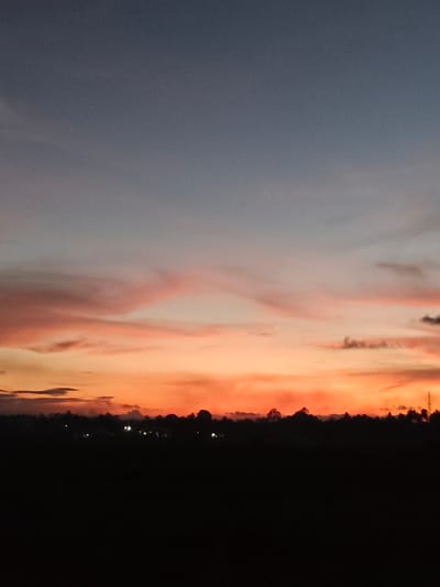 The image depicts a silhouetted landscape against a vibrant sunset. The sky transitions from a deep blue at the top to hues of orange, pink, and yellow closer to the horizon, with scattered clouds adding texture. Below the sky, a dark silhouette of trees and possibly buildings creates a distinct line against the colorful sky. Small, bright lights are visible scattered along the horizon, suggesting a settlement. The foreground is completely dark, obscuring any details on the ground. The overall impression is that of a peaceful evening scene in a rural or semi-rural area.