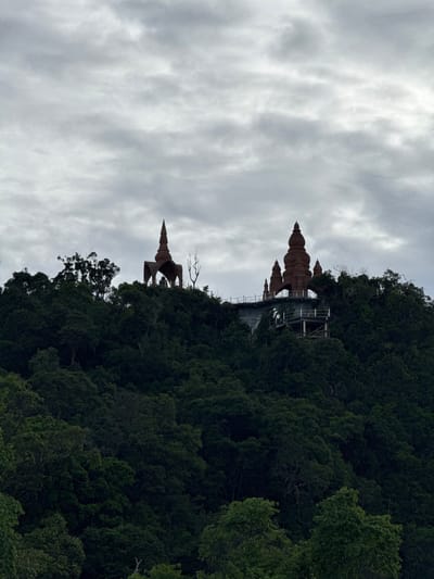 The image captures a view of the Wat Pha Sorn Kaew temple complex, situated atop a lush green hill in Ban Sadet, Thailand. The temple structures, characterized by their unique pagoda-like roofs and rich brown coloring, are silhouetted against a dramatic, cloudy sky. One prominent structure features a large, central pagoda flanked by smaller ones, all appearing to be constructed from mosaic tiles or similar materials. Another structure to the left presents a more open, arched design with a single tall spire. The temples are perched on a constructed platform, evident from the visible railings and supporting structures. The surrounding environment is dominated by dense, dark green foliage, indicating a heavily forested landscape. The overcast weather creates a somber and atmospheric mood. There are no people visible in the image.