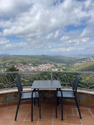 A table and two chairs are set on a tiled patio overlooking a valley with a town nestled within it. The table is black and square, with four black chairs around it. The patio is made of reddish-brown tiles. A decorative metal railing separates the patio from the view. The town, identified as Can Güell, Spain, is spread out below, with buildings featuring red-tiled roofs. The surrounding landscape is covered in lush green trees, and the background shows rolling hills and mountains under a cloudy sky. The scene appears to be an outdoor dining or seating area offering a scenic vista.