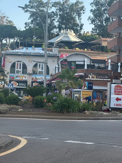 This image captures a street scene in Lloret de Mar, Spain, with a focus on commercial establishments. In the foreground, a section of asphalt road with a yellow curved line and white markings is visible, alongside a concrete curb. Behind the road, a circular planter bed is filled with various plants, including flowering bushes and palm fronds.
Dominating the middle ground are the facades of several businesses. A prominent sign reads "RESTAURANT TERRACE COCKTAIL & SHISHA BAR." Below this, a building features arched windows and large pictorial advertisements for food and drinks, including what appears to be a "BBQ" advertisement. Other businesses visible include "Cocktails," "Chilli Grill," and "LOUNGE." Signs for "Caixer ATM - Cajero" are also present, indicating ATM services.
Further back, the architecture suggests a multi-story urban environment with balconies and brickwork. Trees and foliage are interspersed with the buildings, suggesting a somewhat green urban area. In the background, more buildings and trees can be seen, along with a bright blue sky. A large Burger King sign with a red arrow pointing to the left and the text "1 MIN." is positioned on the right side of the image. The overall impression is a commercial street area, likely a popular spot for dining and entertainment in Lloret de Mar.