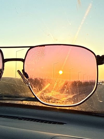 The image shows a close-up of a pair of pink-tinted glasses, held in front of a window. The sun is visible as a bright, yellow-orange orb, appearing to be setting or rising, casting a warm glow across the scene. Light streaks and refractions are prominent, suggesting the sun's rays are passing through the glass of the window and the lenses of the glasses, creating a lens flare effect. The glasses have a black frame and appear to be slightly dirty or smudged, with visible scratches on the right lens.

The background is a soft blur of light and shadow, with vertical lines and textures that hint at an indoor environment, possibly a car interior given the curvature and dark textured elements on the right side of the frame. The overall lighting suggests it is either early morning or late evening. There is no visible text in the image. The scene conveys a sense of warmth and perhaps a moment of reflection, with the focus drawn to the interplay of light and the glasses. The setting is identified as Tankovo, Bulgaria.