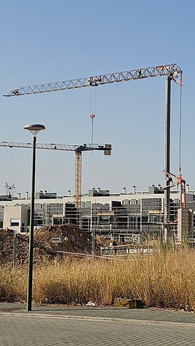 The image depicts a construction site in Cuarte de Huerva, Spain, under a clear blue sky. Dominating the scene are two large construction cranes. The crane on the left is mostly visible, its long horizontal jib stretching across the frame, with a hook suspended from it. To the right, a taller, yellow tower crane is partially obscured by another, larger crane structure. This second, more massive crane has a dark vertical mast and a long jib extending horizontally, with a red component and hook visible. In the foreground, a modern-looking street lamp with a white, rounded light fixture stands on a green pole. Behind the lamp, dry, tall grass fills the mid-ground. Further back, a series of modern apartment buildings, characterized by gray and white facades and dark-paned windows, are under construction. Scaffolding surrounds parts of the buildings, and piles of dirt and construction materials are visible. A small white sign with text is affixed to a fence in front of the dirt piles. The ground in the immediate foreground is paved with gray interlocking pavers.