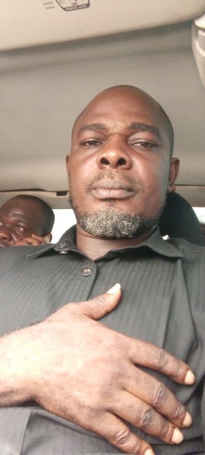 This media file is a close-up, selfie-style portrait of two Black men inside a vehicle. The primary subject, in the foreground, is a bald man with a greying beard, looking directly at the camera with a neutral expression. He is wearing a dark, collared shirt with subtle vertical pinstripes and a decorative button at the collar. His right hand is resting on his chest, with fingers splayed.
Behind him and to the left, another man is partially visible. He has dark hair and is looking towards the left side of the frame, with his right hand near his ear as if holding a phone. The interior of the vehicle is dimly lit, with the ceiling of the car visible at the top of the frame.
At the very top of the image, on the vehicle's ceiling, there is some white text that is partially obscured and difficult to read. The visible portions suggest it might be a label or warning. Based on the provided location context, the scene is likely taking place in Sabon Gari South, Nigeria. There are no obvious cues about the time of day or weather.
