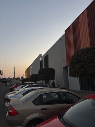 The image depicts several cars parked along a building. The building has a combination of corrugated metal and painted walls, with trimmed bushes lining the front. Two people walk along the sidewalk next to the building. Utility poles extend into the distance, where the sky fades into a hazy sunset. The overall perspective is tilted, giving a slightly disorienting view of the scene.