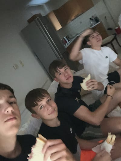 The image shows a group of young people, likely friends, in an indoor setting. One person is lying down wearing a white t-shirt, while three others are positioned closer to the camera. They are all young males, and some are holding pieces of food that appear to be sandwiches. One sandwich is partially eaten and wrapped in white paper. The lighting suggests it might be daytime, but the room appears to be dimly lit, possibly indicating an indoor environment with closed curtains or an evening setting. The overall mood seems casual and relaxed, with the individuals appearing to be enjoying a shared moment, possibly a snack or meal. There is a piece of furniture, possibly a dresser or cabinet, visible in the background. The image is an informal snapshot taken from a low angle, making the subjects appear close and prominent. No visible text is present in the image.