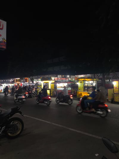 At night, a street scene in Puloara, Indonesia, is captured with motion blur, indicating movement. A row of food stalls with illuminated signs line the right side of the street. One sign reads "KUTARAJA KULINER". Several individuals are riding motorbikes and scooters in the street, some with passengers. The road surface appears wet. The foreground features a parked black motorcycle with a gold rim. The overall atmosphere is that of a busy night market or street food area.