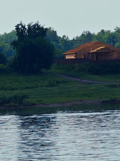 The image depicts a serene natural landscape near Erkalovo, Russia. In the foreground, a body of water with rippling surfaces fills the lower portion of the frame. Beyond the water, a grassy embankment slopes upwards, dotted with patches of greenery. Further inland, a dirt path winds through the grass. Dominating the middle ground is a partially constructed wooden house, its roof unfinished and some framing exposed. Behind the house, a dense line of trees forms a dark green backdrop against a pale, overcast sky. A solitary, dark-leaved tree stands prominently to the left of the house, its branches reaching upwards. The overall scene suggests a rural or semi-rural setting, possibly during the daytime under diffused light. No people or animals are visible.