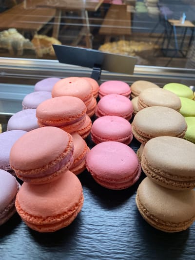 A close-up, eye-level shot showcases a tiered display of colorful macarons on a dark surface, likely a bakery counter in Grainau, Germany. The macarons are arranged in stacks and rows, with visible layers of filling between the cookie halves. Colors include pastel purple, pink, peach, light brown, and light green. The background is blurred, showing a display case with baked goods and reflections of interior lighting and possibly a window. The lighting is soft, highlighting the texture and sheen of the macarons. No people are visible in the frame.