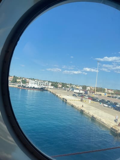 The image, taken from the interior of a vessel through a circular porthole, displays a bright, sunny day at a port. The foreground is dominated by the clear blue water of the sea, reflecting the sky. Beyond the water is a concrete pier lined with parked cars and a few people. Several vehicles, including sedans and SUVs, are visible, suggesting a ferry terminal or a busy port area. A large ship, possibly a ferry or cargo vessel, is docked to the left, with "ALEXANDR" visible on its side. In the background, a town with buildings and greenery can be seen stretching along the coastline. A tall lamppost stands near the pier. The sky is a vibrant blue with scattered white clouds. The location is the Municipal Unit of Kastro - Kyllini, Greece, a port known for its ferry connections to the Ionian Islands.