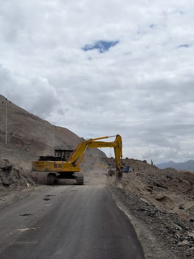 A wide-angle, eye-level shot captures a construction scene on a road in Panamik, India, under a cloudy sky. A large yellow excavator, identified by the "TATA HITACHI" logo, is actively engaged in moving dirt from a rocky mountainside onto the road. The excavator's arm is extended, and its bucket is scooping material, creating a dust cloud. To the right of the excavator, a blue truck is parked, likely to receive the excavated material. Several individuals, appearing as small figures in the distance, are present near the truck and the construction site, suggesting ongoing work. The road itself is asphalted but shows signs of damage, with several dark patches indicating potholes. The surrounding environment is rugged and arid, with a steep, rocky mountain slope dominating the left side of the frame and a pile of excavated debris on the right. In the far background, snow-capped mountains are visible against the horizon. The scene depicts an active road construction or maintenance project in a mountainous, remote area.