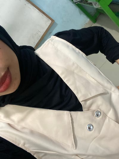 The image shows a close-up of a person wearing a black hijab and a light-colored vest with two decorative gem-like buttons. The person's lips are visible, painted with red lipstick. In the background, a white board with a wooden frame and a light blue wall are visible. A green metal structure, possibly a table or shelf, is also present in the background. The overall lighting suggests an indoor setting. The location is in the city of Tanjung Gading, Indonesia.