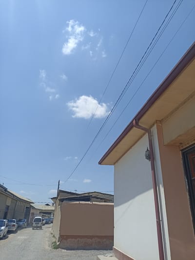 The image depicts a street scene in Chust, Uzbekistan, under a clear blue sky with scattered clouds. The foreground is dominated by the right side of a building with a white and tan facade, featuring a dark brown gutter system and decorative trim. Several power lines crisscross the sky above the street. The street itself is unpaved, with a gravelly surface. On the left side of the street, several cars are parked and in motion. The buildings lining the street appear to be residential or commercial structures with corrugated metal roofs. The overall impression is of a typical day in a moderately developed urban area.