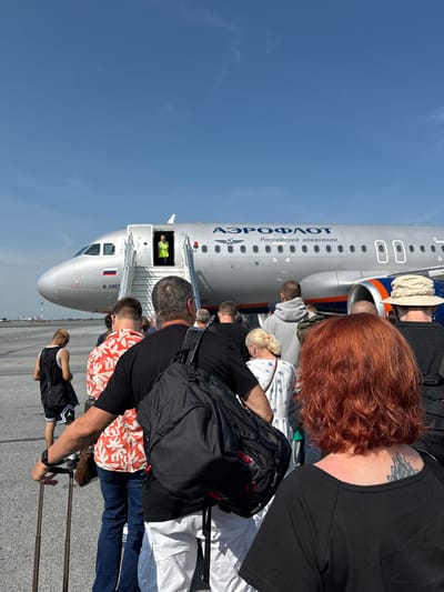 In Kalinino, Russia, a queue of passengers, some carrying luggage, are boarding an Aeroflot airplane. The aircraft, a silver passenger jet with "AEROFLOT" and "Российские авиалинии" written on its fuselage, is parked on the tarmac under a clear blue sky. A worker in a high-visibility vest stands at the open aircraft door, guiding passengers up the stairs. The foreground is filled with the backs of several individuals, their clothing diverse in color and style. One man in the front is holding the handle of a rolling suitcase. The overall scene captures the routine process of air travel.