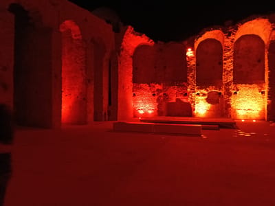 This is a nighttime, long-exposure photograph of the interior of the Nuraghe Palmavera ruins in Alghero, Sardinia, Italy. The ancient stone structure is illuminated with intense red and orange lights, casting long shadows and highlighting the weathered texture of the masonry. The scene is empty of people. The floor appears to be sandy or dirt, with some debris or puddles reflecting the light. Two rectangular, light-colored structures, possibly display plinths or seating, are visible in the foreground, one partially submerged in the illuminated area. The ruins feature arched doorways and rough-hewn stone walls, indicative of ancient Nuragic architecture. The black sky serves as a stark backdrop to the illuminated ruins.