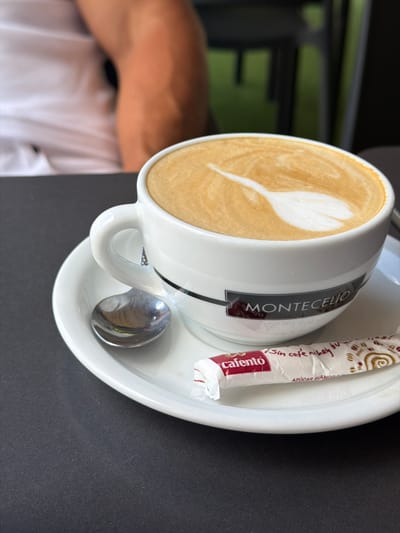A white cup and saucer hold a latte with heart-shaped foam art and a packet of "Cafento" sugar. The cup features a black band with the white text "MONTECELIO". A shiny silver spoon rests on the saucer. The scene is set on a dark gray table, with a blurred background showing a person in a white shirt and an arm. The lighting suggests daytime, and the presence of a cafe setting implies an indoor or outdoor seating area. The overall mood is relaxed and inviting, characteristic of enjoying a coffee break. The sugar packet also contains Spanish text, including "Sin cafe, ni hoy", and "AZUCAR BLANCO".