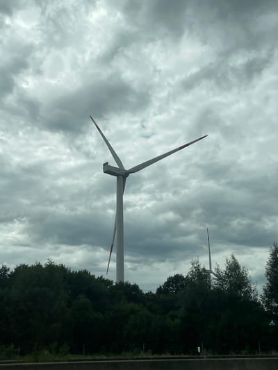 The image depicts a cloudy sky over a treeline with several wind turbines. The primary subject is a large wind turbine in the center of the frame, with its blades extended and its nacelle visible. To the right, a second, smaller wind turbine is partially obscured by trees. A third, even smaller wind turbine is visible in the distance to the far right. The foreground shows a dark treeline, suggesting a rural or semi-rural setting. The overall atmosphere is overcast, with the sky dominated by gray, textured clouds. The scene appears to be taken from a road, with a dark barrier or road surface visible at the bottom of the frame. The specific location is identified as the municipality of Großenaspe, Germany.