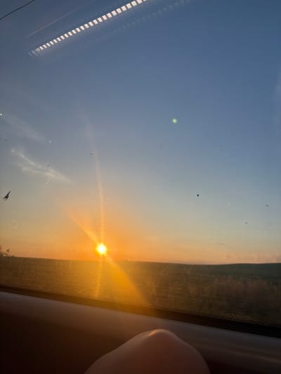 The image captures a sunset viewed from inside a vehicle, likely a train or bus, as evidenced by the reflection of interior lights and the window frame. The sun is low on the horizon, casting a warm, golden glow across the sky and the landscape. The sky transitions from a pale blue at the top to shades of orange and yellow near the horizon. In the foreground, a portion of a person's hand is visible, resting on the window sill. The exterior scene shows a field or rolling hills, with the detail somewhat obscured by the sunset's glare and the window's condition, which has some smudges and debris. The location of Tsarevtsi, Bulgaria, is not visually represented or discernible from the image content.