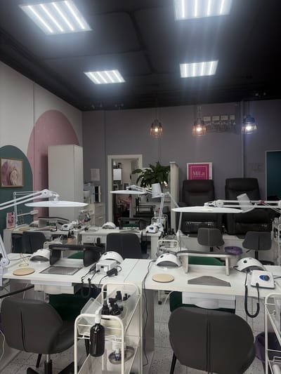 The image depicts the interior of a nail salon in Romanovka, Russia. The scene is set with multiple nail stations, each equipped with white tables, adjustable lamps, nail drills, and chairs for clients and technicians. Several dark, plush chairs are visible, suggesting a comfortable environment for clients. On the tables, various nail care products and tools are neatly arranged, including bottles of polish, brushes, and manicure bowls. A metal rolling cart filled with supplies is also present.
In the background, there are elements of decor such as a framed picture of a flower and abstract wall art featuring pink and green circles. Lighting fixtures are strategically placed throughout the salon, including rectangular fluorescent lights on the ceiling and industrial-style pendant lamps. The overall atmosphere appears clean, organized, and professional, designed to provide nail services. The image is well-lit, suggesting it was taken during daytime business hours. There are no people visible in the immediate foreground or actively engaged in services, but the setup clearly indicates the salon is operational or ready for business. Visible text includes "MiLa" and "Салон красоты" on a pink sign, translating to "Beauty Salon".