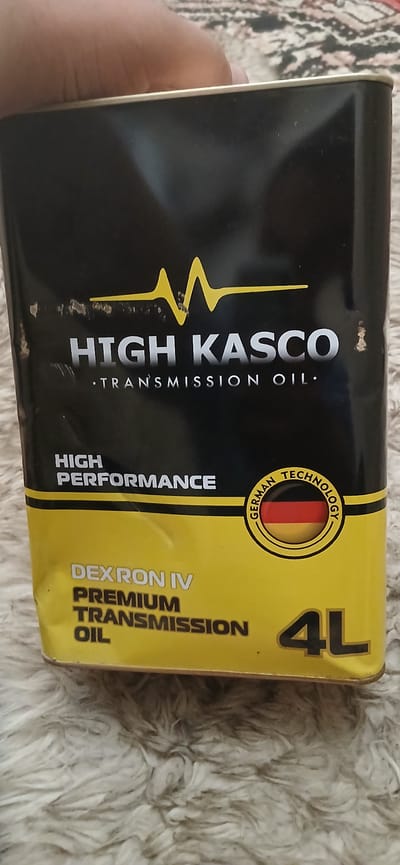 The image shows a black and yellow rectangular metal container of "HIGH KASCO TRANSMISSION OIL". The container is oriented vertically, with a yellow graphic line resembling a sound wave at the top. The brand name "HIGH KASCO" is displayed prominently in large white letters, followed by "TRANSMISSION OIL" in smaller white letters. Below this, "HIGH PERFORMANCE" is written in white. To the right, a circular emblem with the German flag and the text "GERMAN TECHNOLOGY" is visible. The lower half of the container is yellow and displays "DEXRON IV PREMIUM TRANSMISSION OIL" in white and black letters, along with "4L" in large black numerals. The container has some minor scuffs and dents. The container is resting on a fuzzy, beige-colored surface. A person's hand is partially visible at the top of the image, suggesting the container is being held or is on a surface near a person. There is no indication of the city of Kasansay, Uzbekistan, in the image.