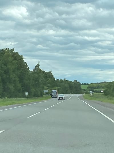 A vertical, eye-level shot captures a two-lane highway in rural Russia, under a cloudy sky. The road stretches into the distance, with white dashed lines marking the lanes. To the left, a dense forest of green trees lines the highway. On the right side, the landscape transitions to rolling green hills with fields in the distance. A silver car is traveling in the left lane ahead, and a large truck is in the right lane further up the road. A few traffic signs are visible on the side of the road. The overall scene suggests a quiet, overcast day in the countryside.