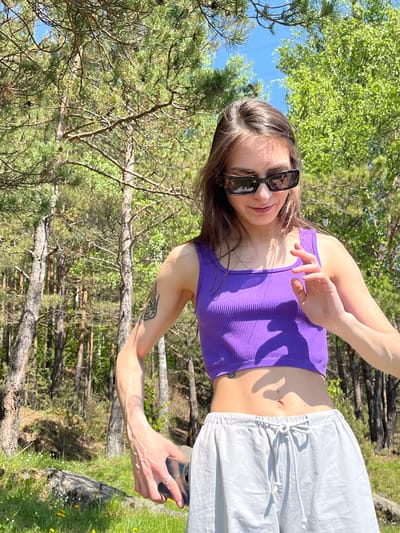 A young woman is captured in an outdoor setting in Слизнево, Russia, appearing to take a selfie or record a video with her smartphone. She is positioned slightly angled, with her head and torso rotated towards the camera, and her right arm extended forward holding the phone. She wears dark rectangular sunglasses, a ribbed purple crop top, and light grey or white drawstring pants. A silver chain necklace is visible around her neck. A small tattoo in the shape of the Venus symbol with an additional horizontal line is visible near her navel, and another tattoo, possibly depicting wings, is on her left forearm. A silver ring adorns her right ring finger.
The background is a vibrant forest or wooded area, dominated by tall pine trees and some deciduous trees with lush green foliage, suggesting spring or summer. The sky above is clear and blue. Bright sunlight casts distinct shadows, indicating a sunny day, likely during the mid-day hours. The ground is covered with green grass, and small yellow and white wildflowers are visible in the foreground to the right. Several fallen tree trunks and branches are scattered throughout the scene. The woman has a slight, relaxed smile. No discernible text is visible on her clothing or the immediate surroundings.