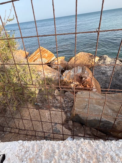 The image shows a rocky shoreline with the Mediterranean Sea in the background. A rusty metal fence runs horizontally across the frame, partially obscuring the view. Large, rough-hewn rocks, some with streaks of orange and white, are scattered on the shore. Green, dry brush grows between the rocks. The water is a deep blue, with small waves. The sky is clear and pale blue. The scene is located in Urbanización Cabo Roig, Spain. The overall impression is one of a rugged, natural coastal environment.