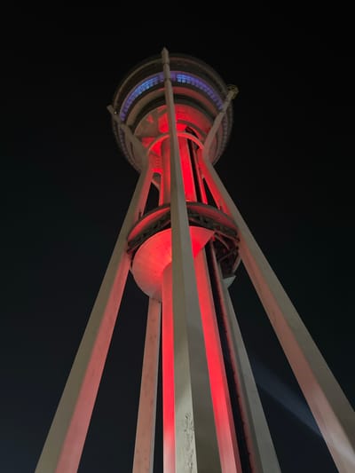 The image shows a low-angle, upward view of the Alor Setar Tower (Menara Alor Setar) at night in Alor Setar, Malaysia. The tower is illuminated with vibrant red lights that shine up its central structure and along its exterior supports. The top section of the tower features blue and purple lighting. The sky is dark, indicating nighttime. The tower's concrete legs are visible extending upwards, supporting a multi-tiered structure at the top. The overall impression is one of architectural grandeur and modern illumination against the night sky.