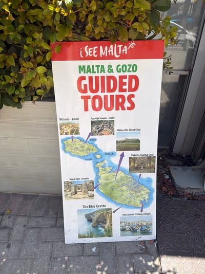 This is a photograph of a sign advertising "iSEE MALTA MALTA & GOZO GUIDED TOURS." The sign is predominantly white with large red and green text. It features a map of Malta and Gozo with various points of interest highlighted and illustrated with small photos.
The illustrated locations include:
- Victoria - Gozo
- Ggantija Temples - Gozo
- Mdina (the Silent City)
- Valletta (Capital City)
- Comino
- Hagar Qim Temples
- The Blue Grotto
- Marsaxlokk (Fishing Village)
The sign is positioned outdoors, partially obscured by lush green foliage. The background shows a building facade and a paved ground. The lighting suggests it is daytime, possibly sunny with dappled shadows on the ground, indicating an outdoor setting. The overall impression is promotional, aiming to attract tourists to explore these notable sites. The text is clear and legible.
