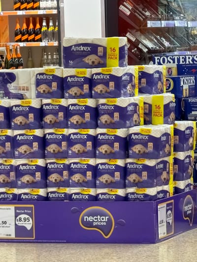 The image shows a stack of Andrex Ultimate Quilts toilet paper, specifically the 16-roll family pack, displayed in a store. The packs are stacked on top of each other, forming a pyramid-like arrangement. A blue "Nectar Prices" promotional box sits beside the toilet paper, indicating a discounted price of £8.95 (compared to the original price of £11.50). The location of the store is suggested by the "Nectar Price" label, which is associated with the Sainsbury's supermarket chain, which may potentially be situated in Pity Me, United Kingdom. The packs of toilet paper feature the Andrex brand name in bold letters and an image of a Labrador puppy. A shelf displaying bottles of alcoholic beverages like Prosecco is located to the left of the toilet paper display. Another brand of lager, Fosters, can be seen in the upper left part of the image.