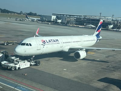 This image depicts a LATAM Airbus A321 aircraft, registration PR-XBG, parked on a concrete tarmac at São Paulo/Guarulhos – Governor André Franco Montoro International Airport (SBGR) in Guarulhos, Brazil. The aircraft, predominantly white with a blue and red LATAM livery on its fuselage and tail fin, is connected to a white WFS pushback tug (marked TL008) at its nose landing gear, indicating ground handling operations. Rows of passenger windows are visible along the fuselage, and one turbofan engine is prominent under the wing on the left side of the aircraft.

In the mid-ground, another LATAM aircraft, identifiable as an Airbus A320 family member, is parked at a gate and connected to a jet bridge. Various ground support equipment, including baggage carts and smaller service vehicles, are positioned around the distant aircraft on the tarmac. The background features a large, multi-story airport terminal structure under apparent construction or renovation, exhibiting exposed concrete and steel frameworks, with multiple jet bridges attached. A treeline borders the far perimeter of the airport, under a clear, light blue sky. Strong shadows cast by the aircraft and ground equipment suggest bright daylight conditions. No discernible people are individually visible in the scene.