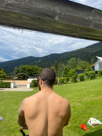The image depicts a person from the back, facing a scenic landscape with a volleyball court and a building in the background, set against a backdrop of lush green hills and a cloudy sky. The person is shirtless and appears to be wearing sunglasses. In the foreground, a wooden beam with mossy growth runs horizontally, partially obscuring the sky. The environment suggests an outdoor recreational area, possibly a park or sports facility.
The precise location of the image is the Oberammergau Sports and Leisure Centre in Oberammergau, Germany. The facility features a beach volleyball court, a children's playground, and a swimming pool, all surrounded by well-maintained lawns and trees. The building visible in the background is likely part of the sports complex. The overall impression is one of a sunny day in a recreational setting, with mountains and forests characteristic of the Bavarian Alps in the distance.