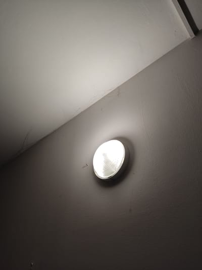 An interior shot captures a circular light fixture mounted on a textured, dark grey wall. The light fixture emits a bright, white glow, illuminating the surrounding area. Above the wall, a white ceiling meets the wall at an angle, showing subtle variations in texture and some faint lines or marks. The overall lighting is dim, with the brightest point being the light fixture itself. The scene appears to be a corner of a room or hallway. No people or specific city landmarks are visible.