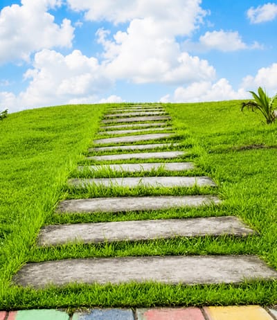 An upward-angled shot captures a stone pathway leading up a grassy hill under a bright blue sky filled with fluffy white clouds. The pathway is made of evenly spaced, rectangular grey stone slabs embedded within lush green grass. The grass is neatly trimmed, with some blades growing between the stones. In the background, the hill slopes upwards and disappears into the sky. To the right, a small, stylized plant with dark green leaves emerges from the grass. At the very bottom of the image, a section of multi-colored paving stones is visible, featuring red, blue, green, and yellow. The scene depicts an outdoor environment, possibly a park or garden, with a clear and sunny day.