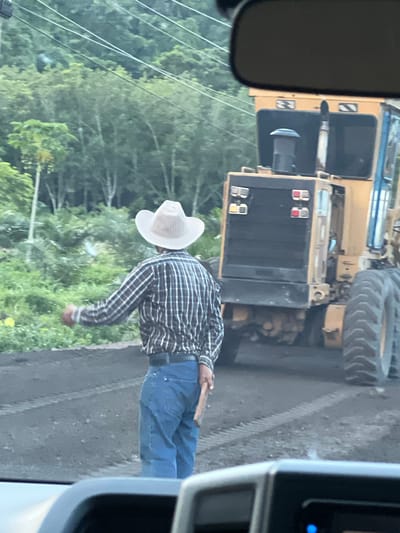 A man wearing a cowboy hat, plaid shirt, and blue jeans stands on a dirt road, gesturing with his right hand while holding a piece of wood in his left. Behind him, a large yellow construction grader is parked. The scene appears to be in a rural area with lush green vegetation and trees in the background. Overhead, power lines are visible. The image is taken from inside a vehicle, as suggested by the rearview mirror and dashboard in the foreground. The exact location in Tambon Don Sak, Thailand is not explicitly discernible from the image alone, but the presence of construction equipment and natural surroundings are evident.