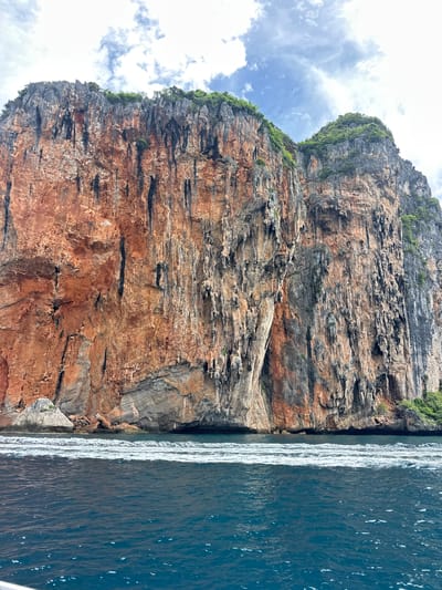 A vast, towering limestone cliff, its face a tapestry of rich red and grey hues, dominates the frame. The cliff is deeply textured, showing signs of erosion with vertical striations and overhangs. Lush green vegetation clings to the summit and parts of the cliff face, indicating a tropical environment. At the base of the cliff, the deep blue water of the sea is visible, with a line of white foam and waves breaking gently. The sky above is partly cloudy, with patches of white cumulus clouds against a pale blue background. The image appears to be taken from a boat, looking towards the shore. The scene captures the dramatic natural beauty of a coastal landscape, likely in a location such as Haad Yao, Thailand, known for its stunning karst formations. There are no people or any discernible man-made objects in the immediate vicinity of the cliff face.